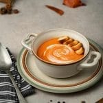 Tasty squash puree soup with fresh cream and toasted bread pieces in bowl on table with black peppercorns on gray background